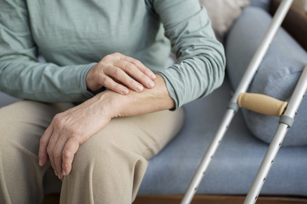 Persona mayor con síntomas de Parkinson sentada en el sofá, apoyando la mano temblorosa sobre la pierna junto a unas muletas.