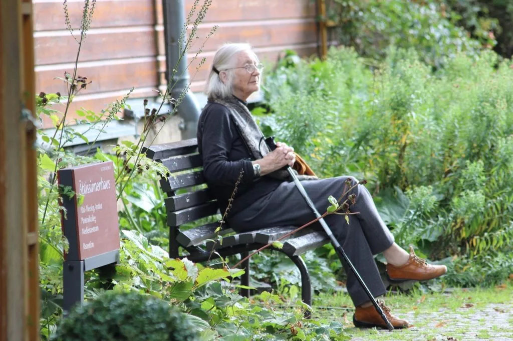 Mujer mayor sentada en un banco al aire libre con bastón, ejemplo de vida sedentaria en la tercera edad.