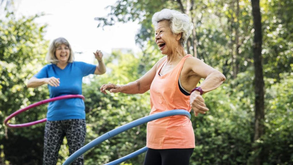 Dos mujeres mayores riendo mientras hacen ejercicio con aros hula hoop al aire libre.