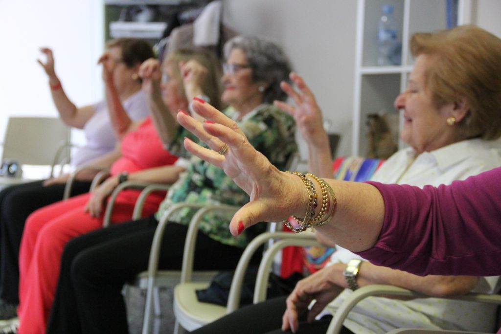 Mujeres mayores sentadas realizando ejercicios físicos adaptados en grupo dentro de una residencia geriátrica.