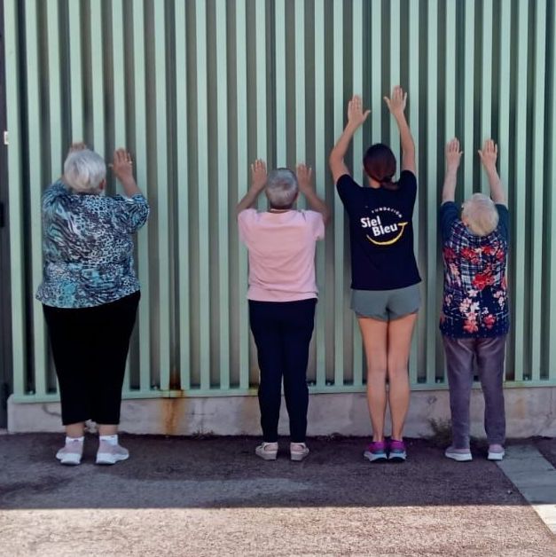 Grupo de personas mayores realizando ejercicios con los brazos elevados apoyados en una pared junto a una entrenadora de Siel Bleu.