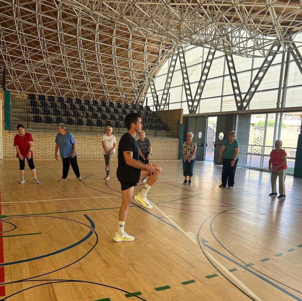 Entrenador guiando a un grupo de personas mayores en una sesión de ejercicio físico adaptado en un polideportivo cubierto.